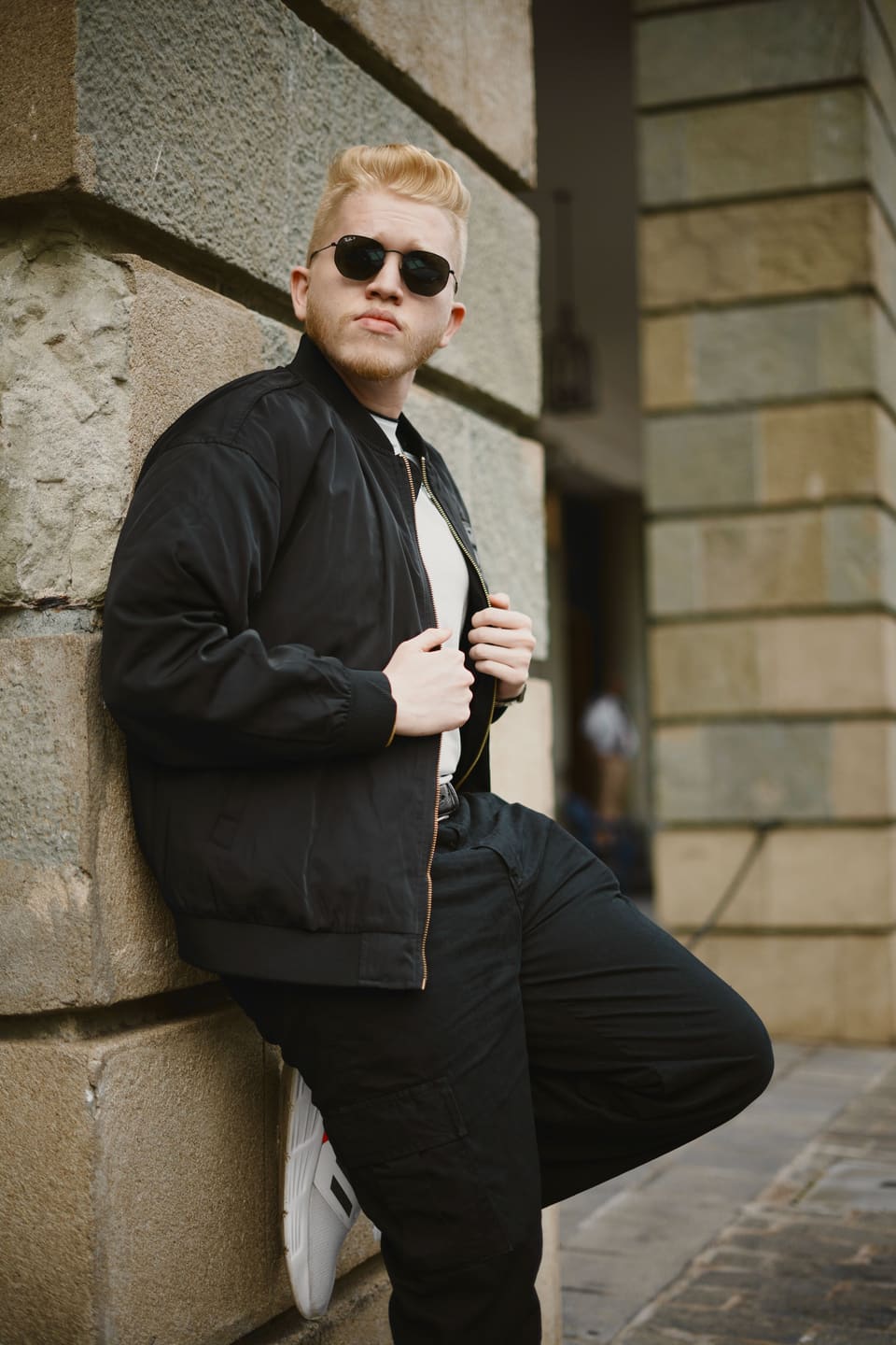 Kenny Dissanayake leaning against a stone wall in a black bomber jacket